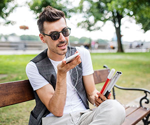 Image of a man wearing sunglasses holding a walking cane and speaking into a smartphone.