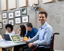 Image of man who uses a wheelchair smiling at camera. He appears in an office setting and is typing on a laptop computer.